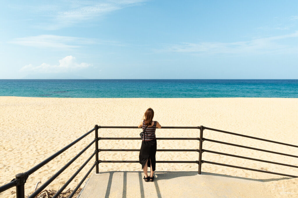 柵に寄りかかって永田浜を見ている女性の後ろ姿