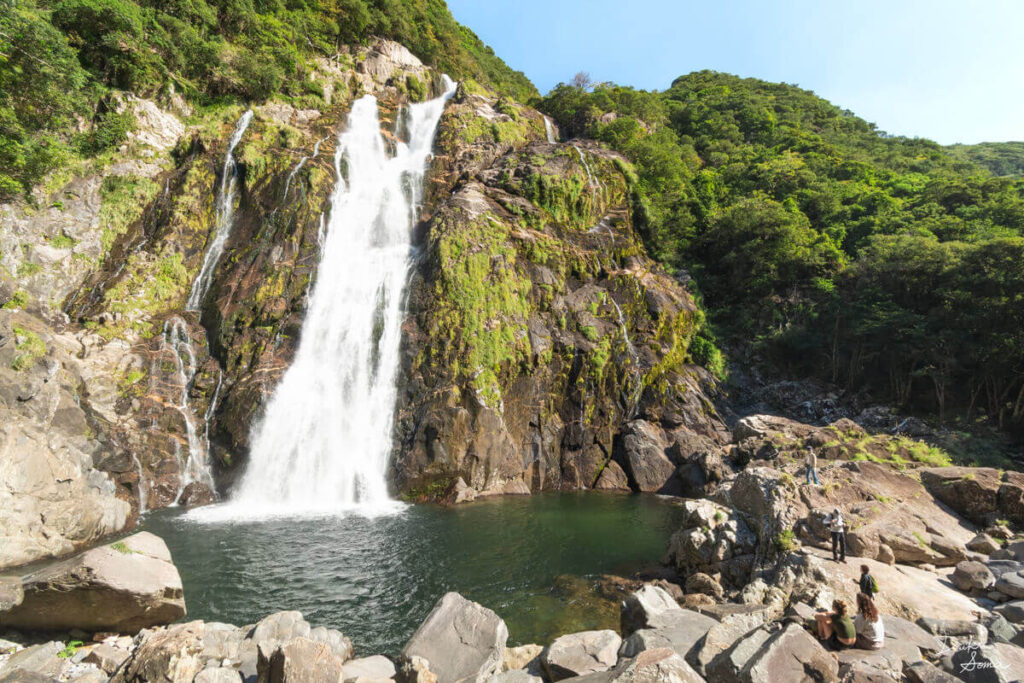 ゴツゴツとした岩壁を流れ落ちる大川の滝と、滝壺の周りで鑑賞する観光客たち